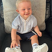 Oliver is registered to the contest to win money with this photo: toddler, child, smiling, sitting, polo_shirt, shorts, white_shoes, socks, leather_chair, cushion, indoor, happy, blonde_hair, face, person, casual_clothing, footwear, young_child, home, comfort