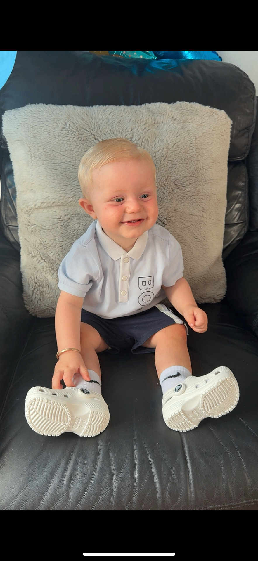 Oliver is registered to the contest to win money with this photo: toddler, child, smiling, sitting, polo_shirt, shorts, white_shoes, socks, leather_chair, cushion, indoor, happy, blonde_hair, face, person, casual_clothing, footwear, young_child, home, comfort