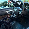 cat, black_cat, animal, pet, feline, sitting, car, car_interior, passenger_seat, steering_wheel, dashboard, leather_seat, luxury_car, door_panel, window, parked_car, storefront, outdoor, portrait, yellow_eyes