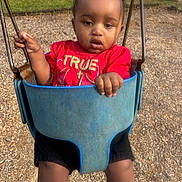 Amir is registered to the contest to win money with this photo: toddler, child, swing, playground, red_shirt, cross_necklace, outdoor, park, wood_chips, grass, stone_wall, daylight, person, baby, hand, legs, metal_chains, seat, casual_clothing, expression