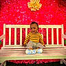 baby, child, bench, smile, yellow_shirt, gray_pants, flower_wall, red_flowers, paper_flowers, white_bench, grass, happy, cute, indoors, portrait, sitting, footwear, colorful, bright, decor