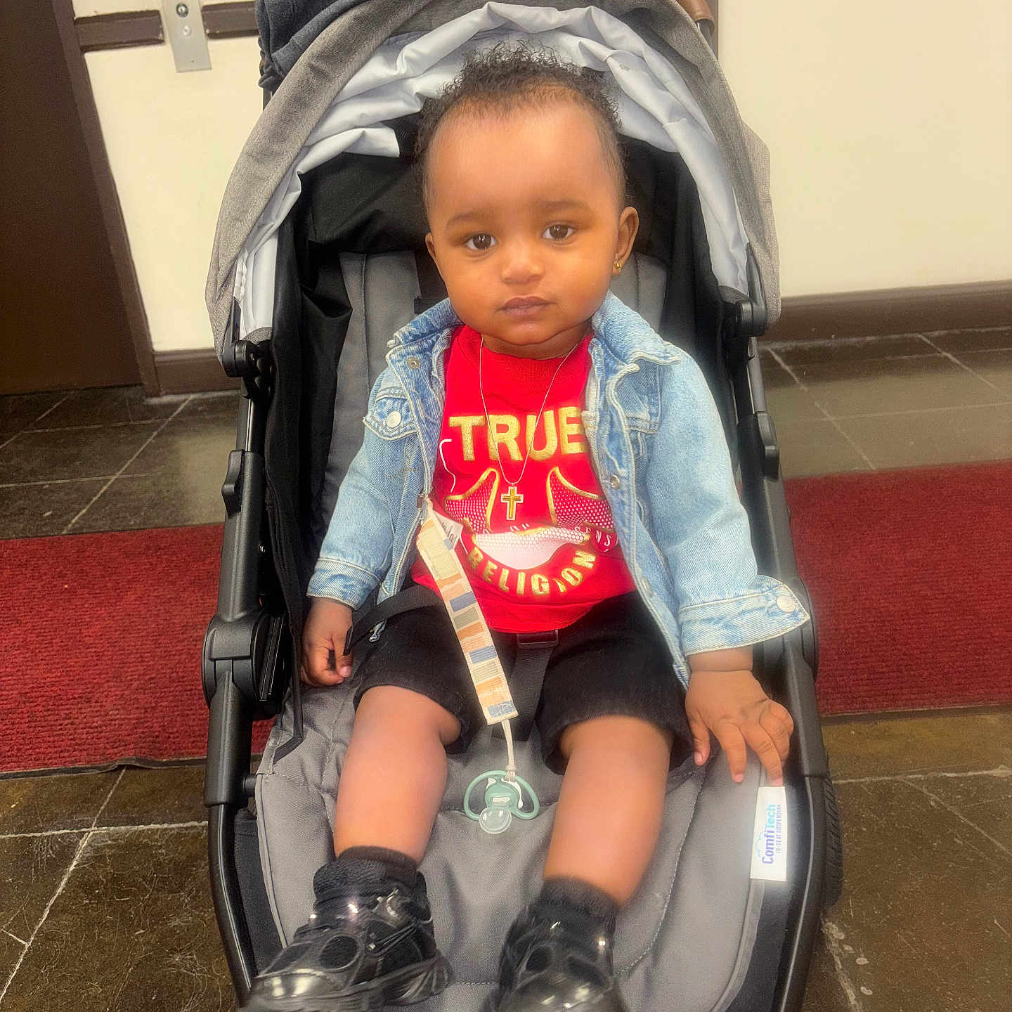 Amir joined the competition — help win amazing prizes! baby, toddler, stroller, child, denim_jacket, red_shirt, black_shoes, pacifier, indoor, floor_tiles, carpet, hand, face, person, clothing, jewelry, earrings, necklace, expression, seated