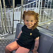 Ylana participe au concours pour gagner de l'argent avec cette photo : child, smiling, curly_hair, red_hair, black_shirt, orange_shorts, tattoo, amusement_ride, metal_bars, seat, night, lights, playful, happy, portrait, cute, fun, person, indoor, young