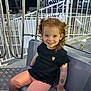 child, smiling, curly_hair, red_hair, black_shirt, orange_shorts, tattoo, amusement_ride, metal_bars, seat, night, lights, playful, happy, portrait, cute, fun, person, indoor, young