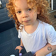 Ylana participe au concours pour gagner de l'argent avec cette photo : child, toddler, curly_hair, white_shirt, tattoo, outdoor, porch, table, wood, plastic, face, person, expression, daylight, casual, young, cute, curious, skin, hand