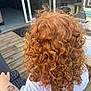 child, curly_hair, red_hair, tattoo, arm, white_shirt, outdoor, wooden_deck, hand, back_view, person, casual, summer, daylight, skin, temporary_tattoo, furniture, door, home, relaxation