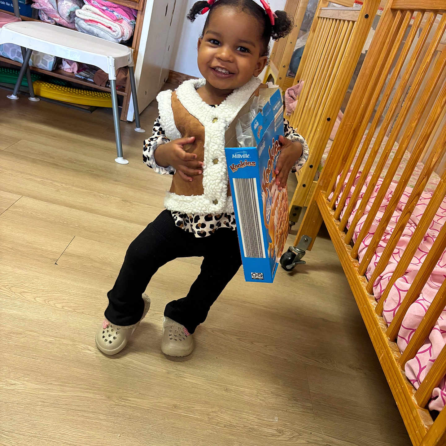 Khloe Knox is registered to the contest to win money with this photo: blanket, cabinet, cereal_box, chair, child, clothing, flooring, hair_bows, happy, indoor, mirror, pants, person, pigtails, playroom, shoes, smiling, toddler, vest, wooden_crib