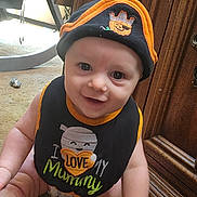 Levi is registered to the contest to win money with this photo: baby, child, smiling, face, hat, bib, indoor, carpet, wooden_cabinet, happy, cute, infant, person, apparel, sitting, closeup, expression, floor, young, portrait