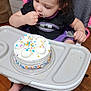 toddler, child, high_chair, cake, frosting, sprinkles, birthday, indoor, wooden_floor, black_shirt, purple_skirt, pink_ruffle, fork, food, curious, sitting, celebration, young_child, person, cute