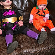 Athena is registered to the contest to win money with this photo: child, costume, superhero, pumpkin, orange, purple, black, mask, couch, leather, indoor, floor, sock, sitting, cute, baby, toddler, bookshelf, decoration, wood_floor