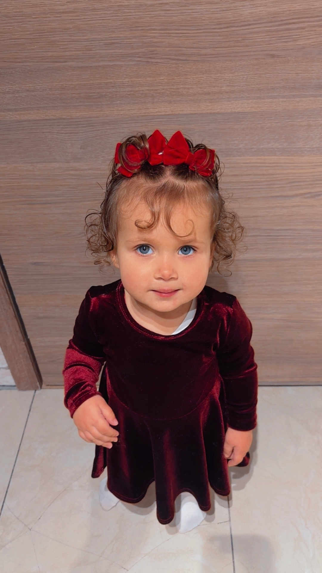 Gabriella a rejoint le concours — aidez-le/la à gagner de superbes lots ! child, toddler, girl, curly_hair, red_bow, blue_eyes, velvet_dress, burgundy, portrait, indoor, wood_background, tile_floor, standing, looking_up, hands, smile, cute, holiday_outfit, closeup, young_child