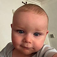 Theodore is registered to the contest to win money with this photo: baby, blue_eyes, child, close_up, curious, cute, expression, face, hair, head, human, indoor, infant, looking_at_camera, onesie, person, portrait, skin, wall, young_child