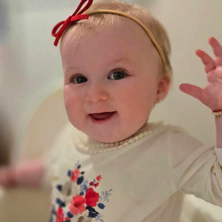 Riki-Hoffman is registered to the contest to win money with this photo: baby, blond_hair, child, closeup, cute, face, floral_pattern, hand, happy, headband, high_chair, indoor, infant, light_skin, person, portrait, red_bow, shirt, smiling, young