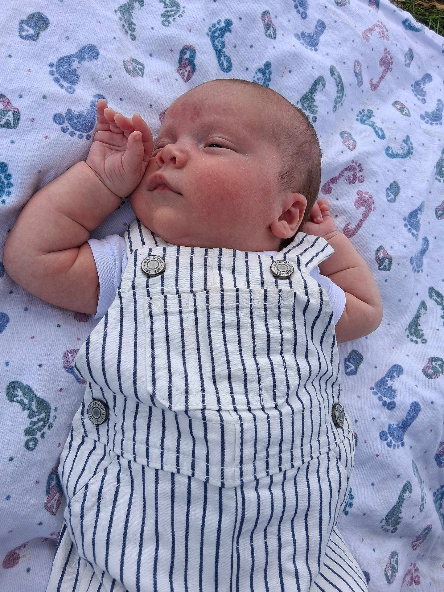 Shane is registered to the contest to win money with this photo: baby, infant, blanket, footprints, striped_overall, clothing, sleepy, peaceful, skin, face, hand, lying_down, outdoor, grass, cute, newborn, portrait, young_child, resting, soft