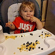 Kai is registered to the contest to win money with this photo: toddler, child, baby, high_chair, cereal, blueberries, food, eating, smiling, happy, red_shirt, indoors, kitchen, tray, messy, portrait, seated, hand, breakfast, playful