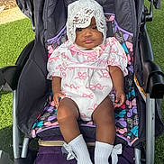 Wynter is registered to the contest to win money with this photo: baby, child, stroller, outdoor, grass, white_outfit, lace_bonnet, pink_bows, socks, infant, cute, portrait, seated, colorful, fabric, sunlight, expression, person, young_child, comfort
