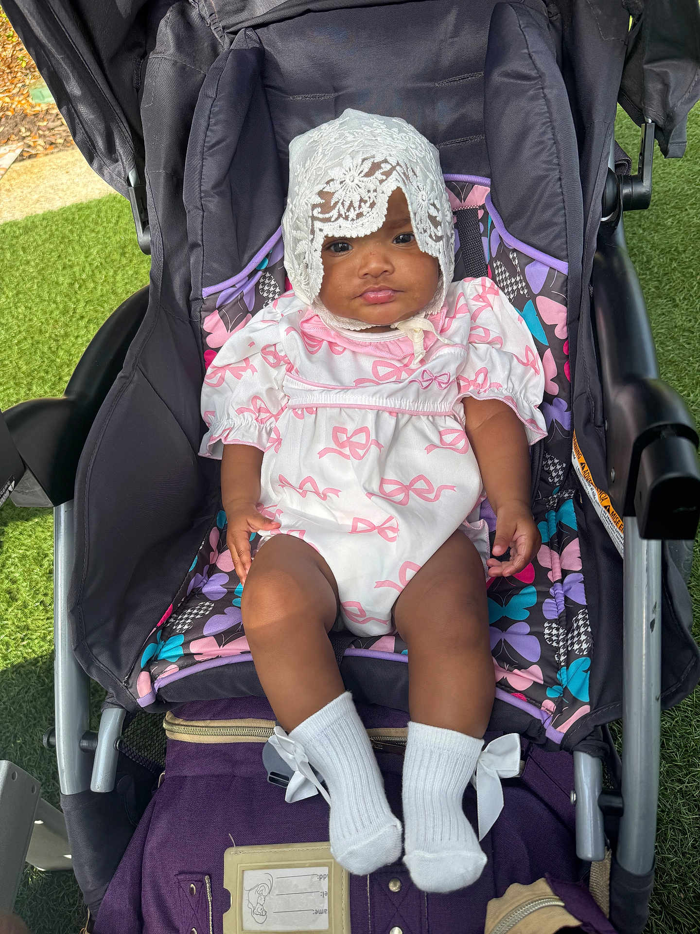 Wynter is registered to the contest to win money with this photo: baby, child, stroller, outdoor, grass, white_outfit, lace_bonnet, pink_bows, socks, infant, cute, portrait, seated, colorful, fabric, sunlight, expression, person, young_child, comfort
