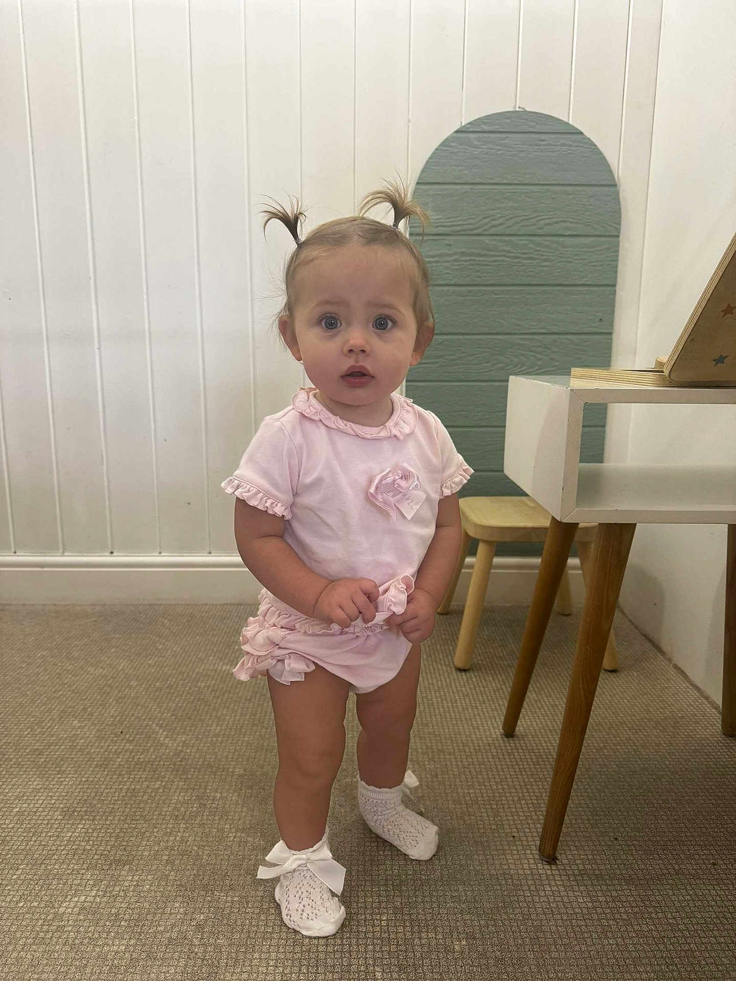 Nellie joined the competition — help win amazing prizes! toddler, child, indoors, pink_clothing, socks, bow, pony_tail, carpet, wooden_chair, table, curious, standing, young_child, cute, frills, baby, person, room, wall_paneling, floor