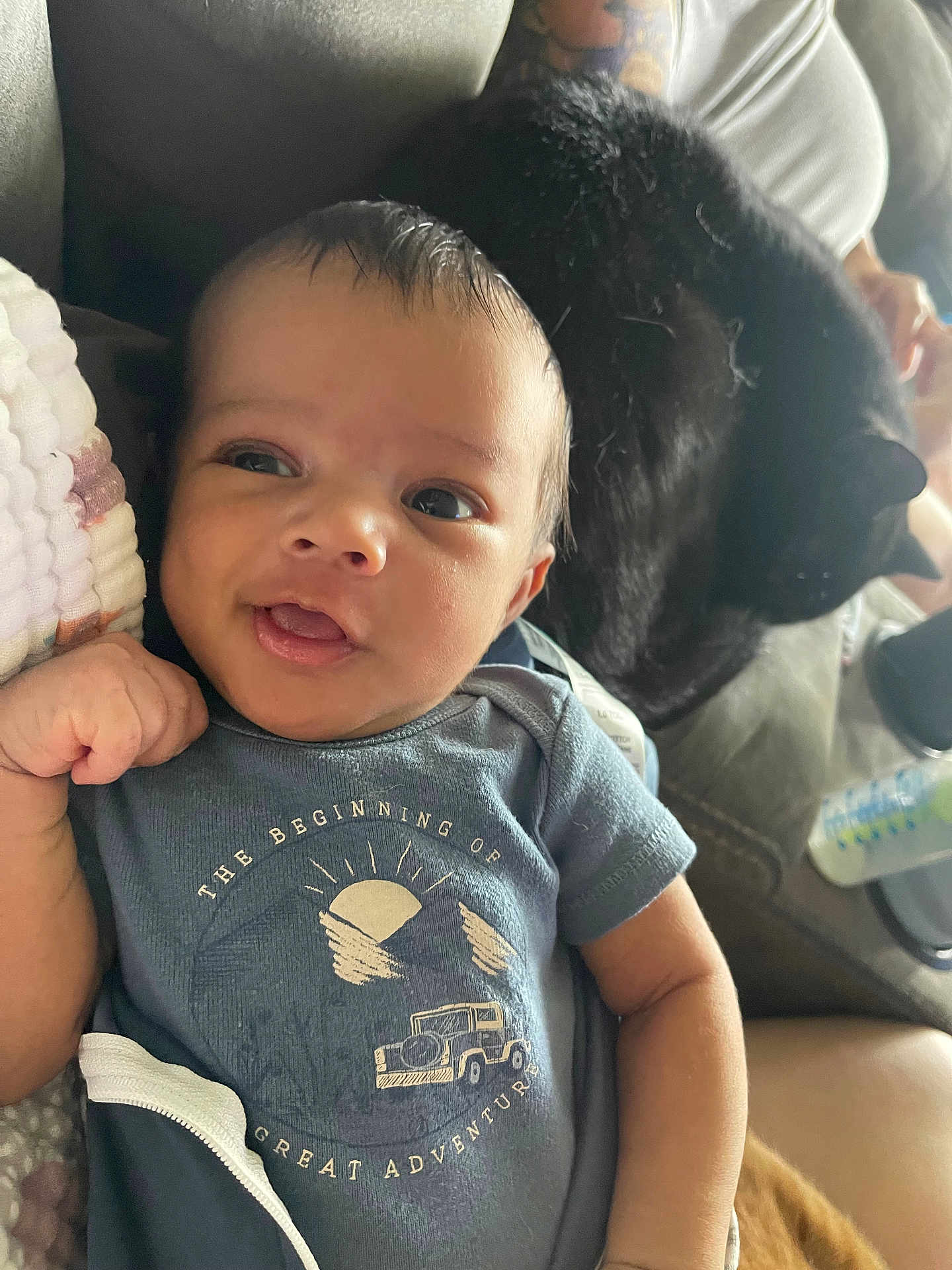 Jensen is registered to the contest to win money with this photo: baby, infant, child, blue_shirt, text_on_clothing, cat, black_cat, pet, couch, indoor, person, cute, smiling, happy, face, hand, relaxing, cozy, home, animal