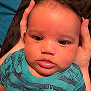 Jensen is registered to the contest to win money with this photo: baby, infant, face, hands, blue_shirt, dinosaur_print, closeup, child, person, skin, eyes, lips, hair, portrait, expression, holding, background, fabric, soft_light, young