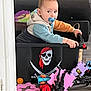 black_toy, blue_toy, casual_clothing, child, container, couch, curious, floor, foam_puzzle, hoodie, indoor, orange_toy, pacifier, person, pirate_skull, plastic_toy, red_toy, seated, toddler, toy