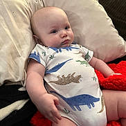 Jasper is registered to the contest to win money with this photo: baby, infant, dinosaur_onesie, couch, pillow, blanket, red_blanket, soft_furniture, indoor, child, cute, relaxed, portrait, person, skin, arm, hand, legs, seated, expression