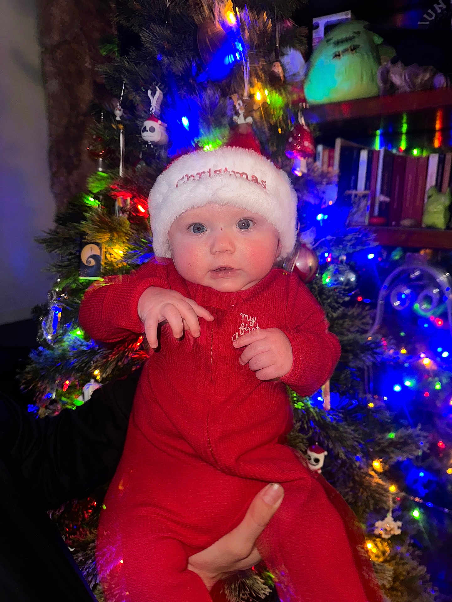 Jasper is registered to the contest to win money with this photo: baby, christmas_hat, red_onesie, holiday, festive, christmas_tree, ornaments, colorful_lights, indoor, child, cute, holding_hand, celebration, smiling, face, person, winter, seasonal, decorations, bright