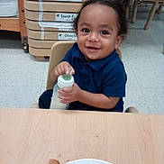 Josiah joined the competition — help win amazing prizes! toddler, child, baby, smiling, portrait, classroom, daycare, seated, bottle, drinking, table, food, plate, storage_bins, furniture, indoor, school, curly_hair, blue_shirt, happy