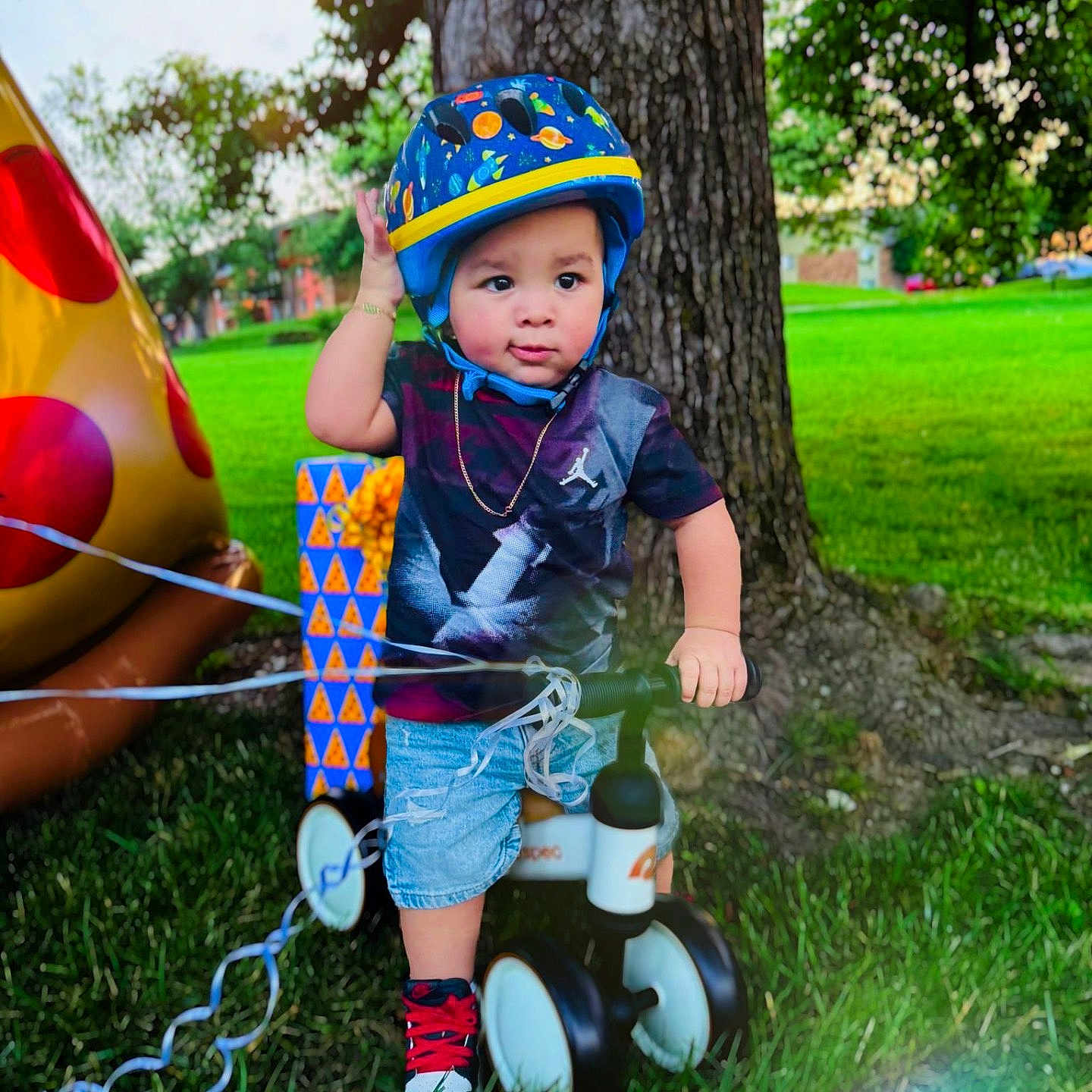 Eithan Dankiel Silva joined the competition — help win amazing prizes! birthday, casual_clothing, child, daylight, gift_box, grass, greenery, helmet, inflatable, outdoor, park, pizza, playing, shorts, sneakers, toddler, toy, tree, tricycle, tshirt