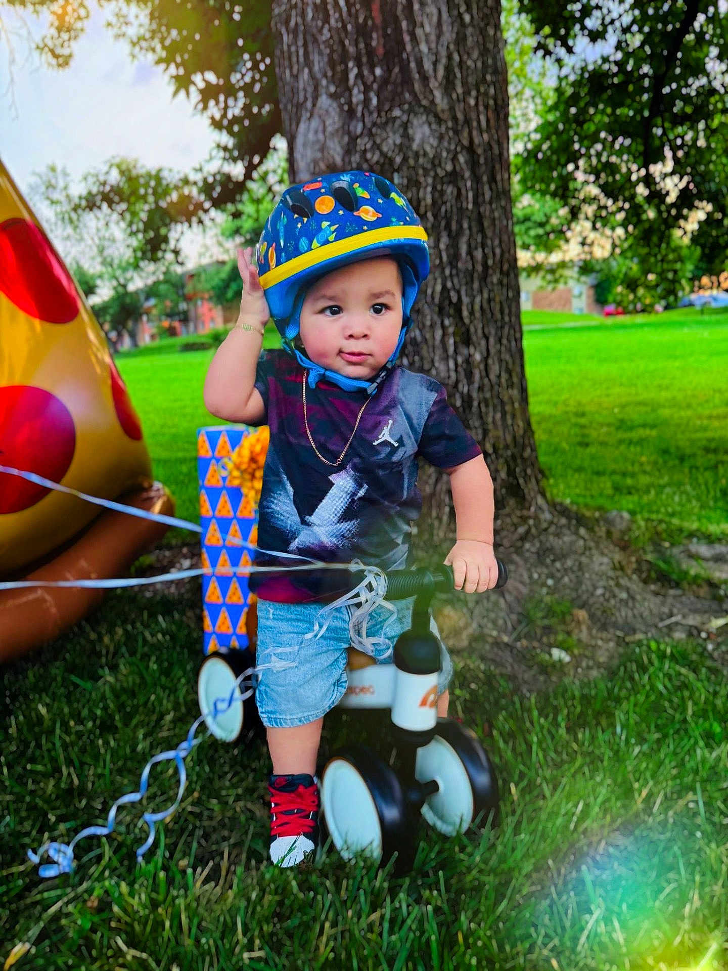 Eithan Dankiel Silva joined the competition — help win amazing prizes! birthday, casual_clothing, child, daylight, gift_box, grass, greenery, helmet, inflatable, outdoor, park, pizza, playing, shorts, sneakers, toddler, toy, tree, tricycle, tshirt