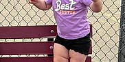 Oaklynne is registered to the contest to win money with this photo: toddler, child, bench, purple_shirt, black_shorts, barefoot, smiling, pigtails, curly_hair, outdoor, chain_link_fence, tennis_court, bicycle, person, car, trees, cloudy_sky, parked_car, asphalt, recreation