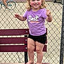 toddler, child, bench, purple_shirt, black_shorts, barefoot, smiling, pigtails, curly_hair, outdoor, chain_link_fence, tennis_court, bicycle, person, car, trees, cloudy_sky, parked_car, asphalt, recreation