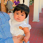 Syeda Fatima joined the competition — help win amazing prizes! baby, child, person, holding, white_dress, striped_shirt, ponytail, curious_expression, brick_walkway, outdoor, storefront, adult, people, seated, red_brick, portrait, cute, infant, young_child, daylight