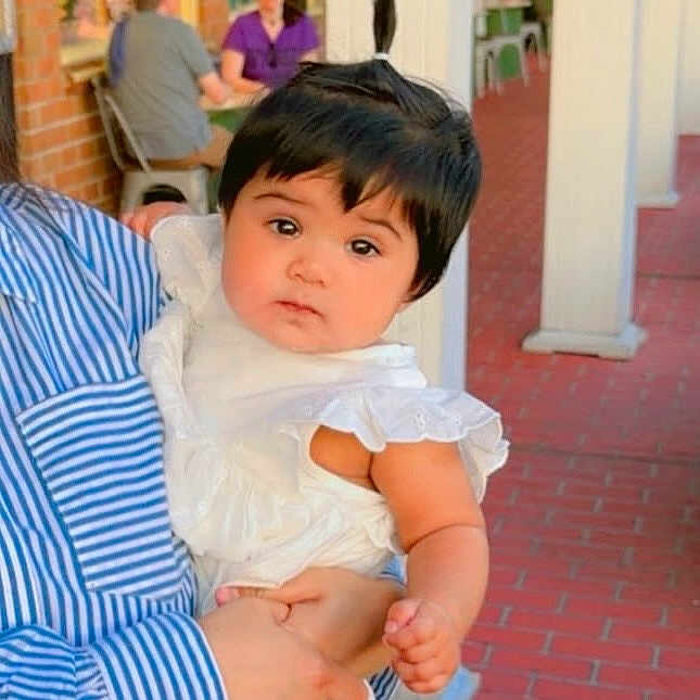 Syeda Fatima joined the competition — help win amazing prizes! baby, child, person, holding, white_dress, striped_shirt, ponytail, curious_expression, brick_walkway, outdoor, storefront, adult, people, seated, red_brick, portrait, cute, infant, young_child, daylight