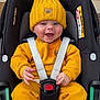 baby, child, car_seat, yellow_clothing, knit_hat, teddy_bear_patch, smiling, happy, infant, straps, sitting, indoor, cute, face, person, seatbelt, safety, clothing, hood, car_seat_harness