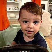 Ryan is registered to the contest to win money with this photo: toddler, child, baby, portrait, face, eyes, brown_hair, smiling, high_chair, indoor, home, play_structure, orange, toy, steering_wheel, shirt, furniture, sitting, background, photograph