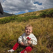 Leyna a rejoint le concours — aidez-le/la à gagner de superbes lots ! baby, child, grass, outdoor, nature, field, clouds, sky, hill, greenery, sitting, cute, sunlight, casual_clothing, red_pants, white_sweatshirt, person, young_child, curly_hair, daytime