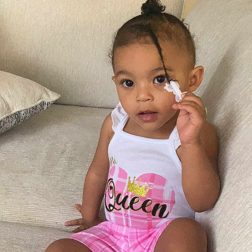 Namirah is registered to the contest to win money with this photo: beige, casual, child, clothing, couch, curly_hair, cute, expression, fashion, hair_clip, home, indoor, person, portrait, relaxed, shorts, sitting, tank_top, toddler, young_child