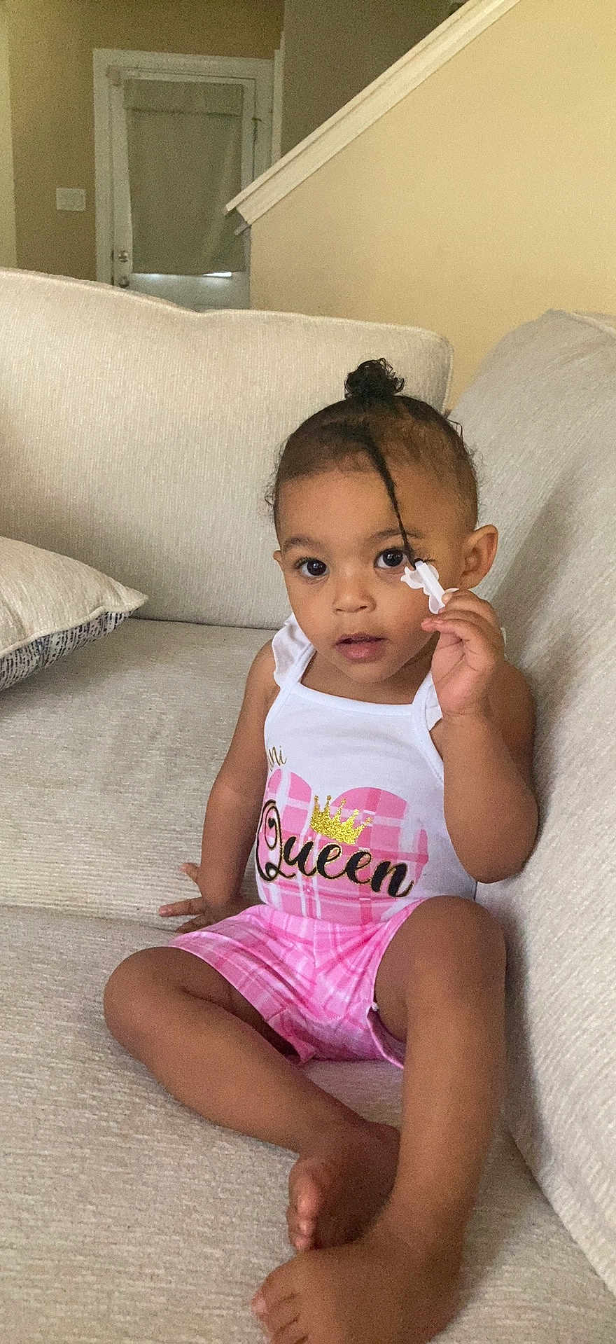 Namirah is registered to the contest to win money with this photo: toddler, child, couch, indoor, clothing, tank_top, shorts, hair_clip, curly_hair, sitting, beige, expression, person, young_child, casual, relaxed, home, cute, fashion, portrait