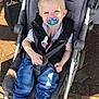 Rio is registered to the contest to win money with this photo: toddler, pacifier, stroller, child, blue_eyes, blond_hair, black_vest, white_shirt, blue_jeans, sneakers, outdoor, sunlight, pavement, baby, seatbelt, footwear, toy_clip, happy, person, casual_clothing