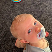 Rio is registered to the contest to win money with this photo: toddler, child, pacifier, blue_eyes, carpet, toy, adult_arm, tattoo, casual, indoor, curious, sitting, clothing, light_hair, person, closeup, playtime, home, floor, looking_up