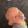 toddler, child, pacifier, blue_eyes, carpet, toy, adult_arm, tattoo, casual, indoor, curious, sitting, clothing, light_hair, person, closeup, playtime, home, floor, looking_up
