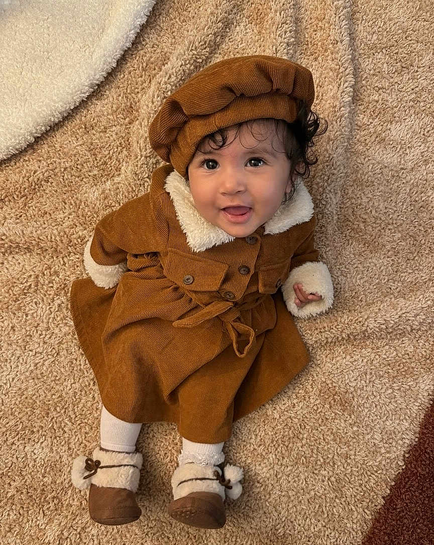 Mirella participe au concours pour gagner de l'argent avec cette photo : baby, child, smile, brown_clothing, hat, carpet, sitting, happy, indoors, footwear, coat, curly_hair, toddler, warm_clothing, cute, portrait, face, person, soft_texture, cozy