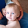 toddler, child, blue_eyes, blonde_hair, face, portrait, skin, person, adult_hand, jeans, carpet, indoor, casual, close_up, candid, family, intimate, soft_light, expression, curious