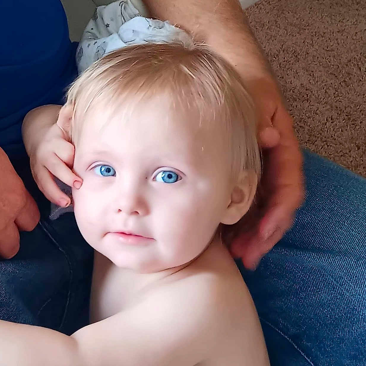 Kayden is registered to the contest to win money with this photo: adult_hand, blonde_hair, blue_eyes, candid, carpet, casual, child, close_up, curious, expression, face, family, indoor, intimate, jeans, person, portrait, skin, soft_light, toddler