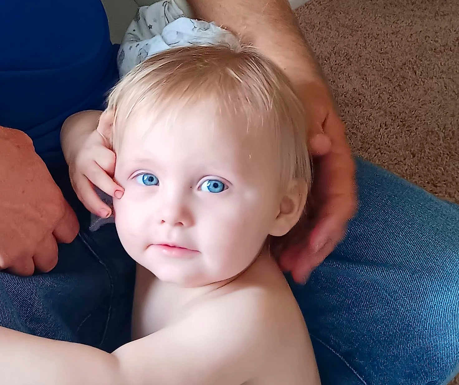Kayden is registered to the contest to win money with this photo: toddler, child, blue_eyes, blonde_hair, face, portrait, skin, person, adult_hand, jeans, carpet, indoor, casual, close_up, candid, family, intimate, soft_light, expression, curious