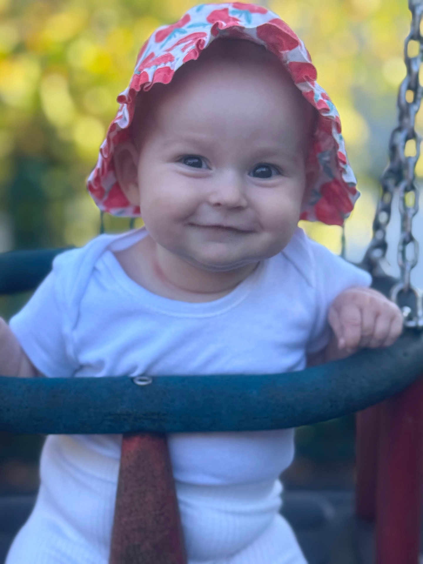 Elowyn is registered to the contest to win money with this photo: baby, child, swing, hat, smile, outdoor, nature, playground, white_clothing, happy, face, person, summer, sun_hat, infant, portrait, hands, seat, blurred_background, daylight