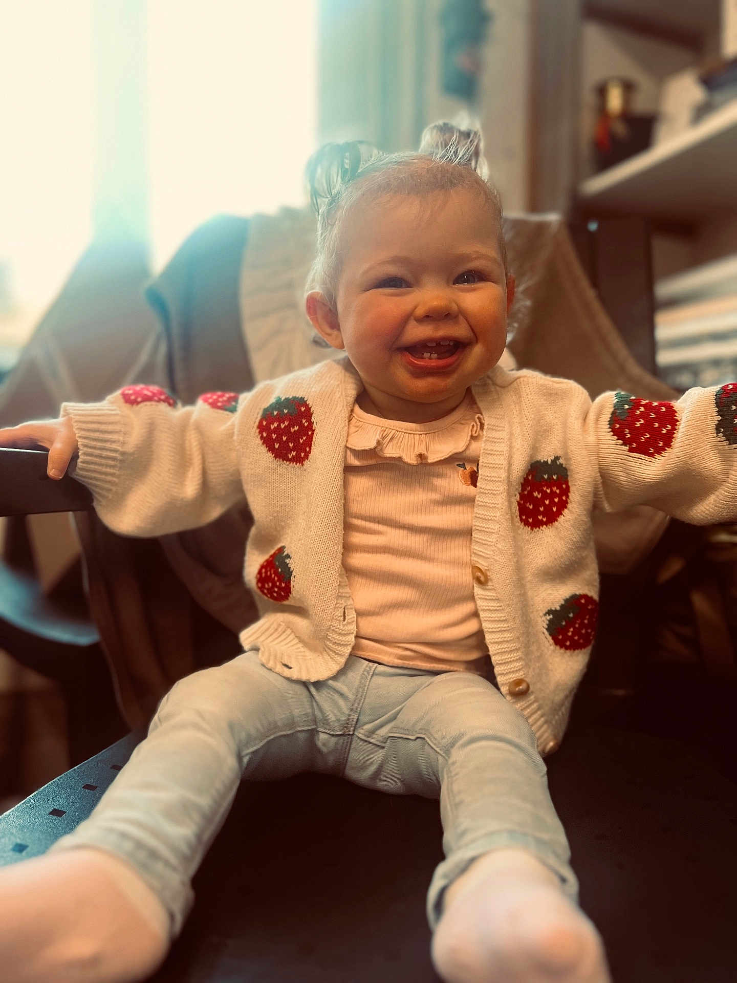Cléa participe au concours pour gagner de l'argent avec cette photo : toddler, child, smiling, sitting, sweater, strawberry_pattern, jeans, sock, indoor, chair, happy, cute, baby, person, portrait, warm_light, cozy, hair_buns, clothing, furniture