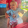 adult, american_flag, baby, backyard, blue_shirt, cartoon_clothing, celebration, child, happy, holding, house, infant, outdoor, parent, patriotic_decorations, red_white_blue, smiling, standing, striped_shorts, sunlight