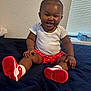 baby, bed, blanket, cheeks, child, cuteness, footwear, hair_tie, happy, indoor, person, polka_dot, red_and_white, red_shorts, sitting, skin, smiling, sneakers, white_shirt, window_blinds
